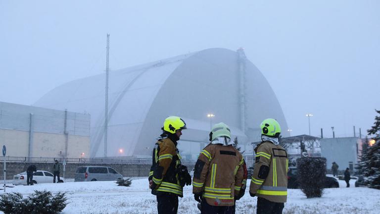 Drei Feuerwehrmänner stehen vor der zum Teil zerstörten Schutzhülle des Kraftwerks in Tschernobyl, welche im Januar 2025 durch eine russische Drohne angegriffen wurde.