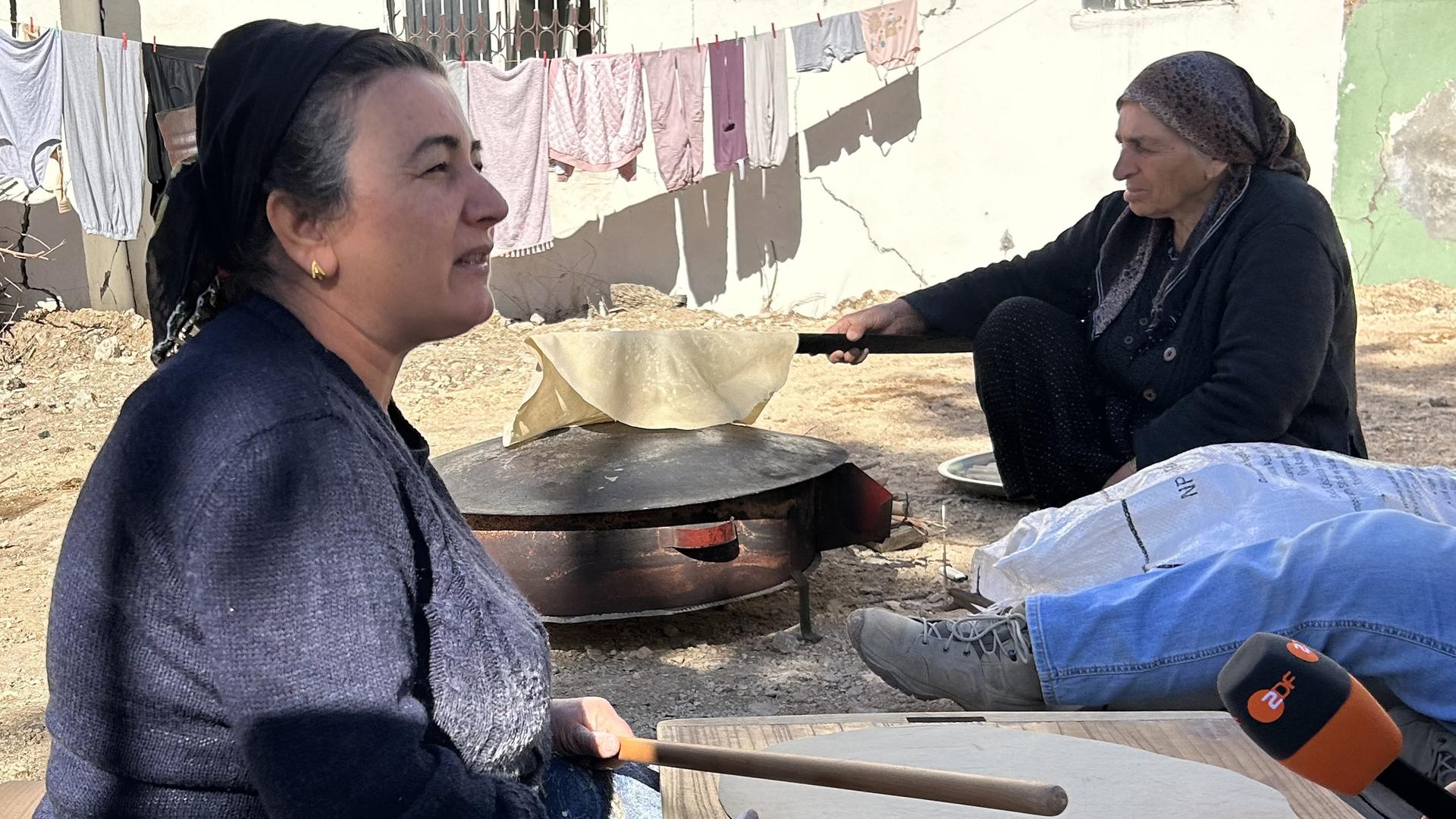 Zwei Frauen sitzen draußen vor einem Haus auf dem Boden und backen Brot