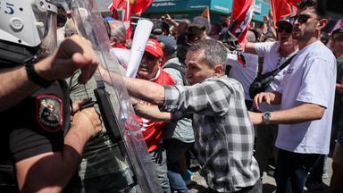 Bei einer Demonstration schiebt ein Mann gegen den Plexiglasschild eines Polizisten in Schutzausrüstung. Hinter ihm schweken Männer rote Fahnen.