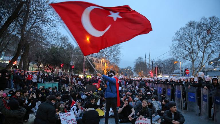 Eine Gruppe an Studenten sitzt mit Protestplakaten in der Hand auf dem Boden, in der Mitte steht ein maskierter Mann, der eine türkische Flagge schwenkt. Auf der rechten Seite befindet sich eine Gruppe Polizisten mit Schutzausrüstung, die die Protestierenden im Zaum hält.