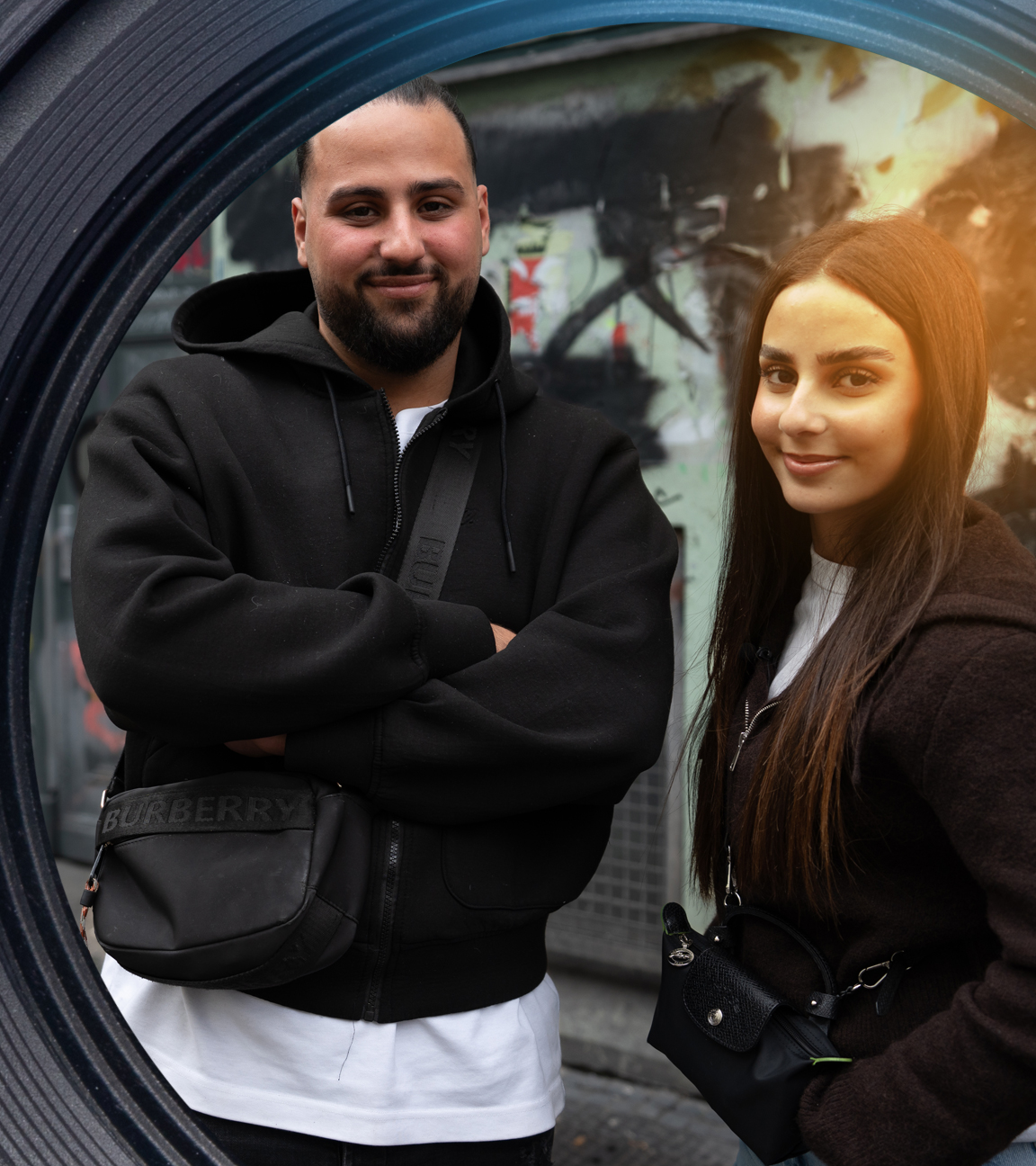 Melih Saydam und seine Schwester Melike stehen auf einer Straße in Berlin. Im Hintergrund ist eine Hauswand mit Graffiti.