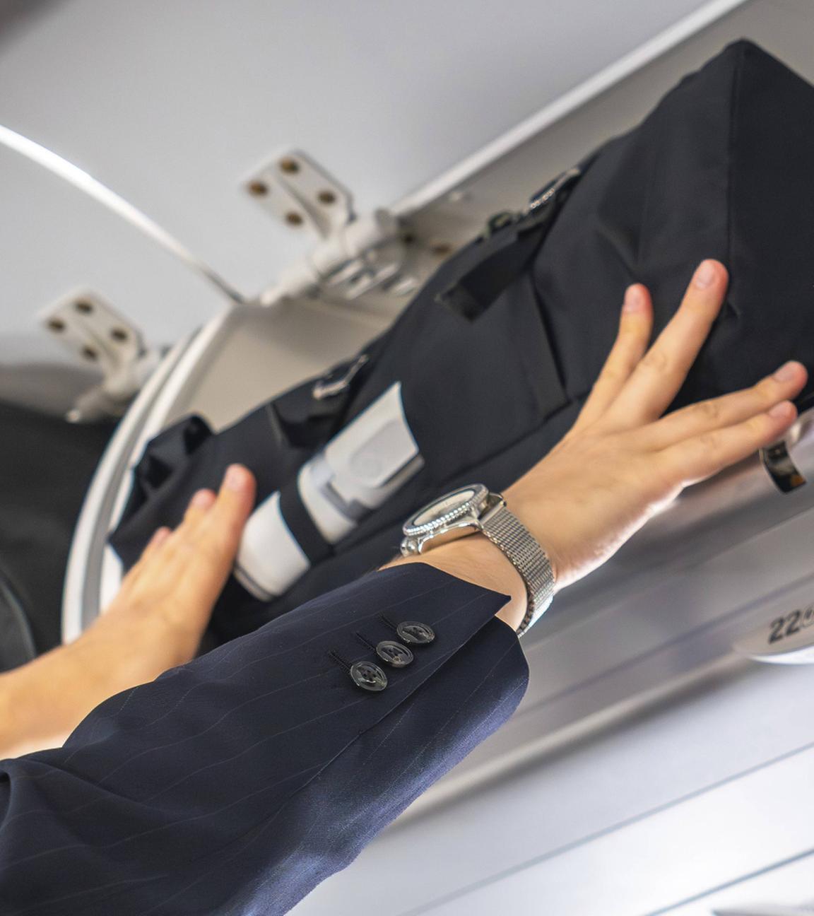 Geschäftsmann mit Anzug und Uhr verstaut während eines Fluges Handgepäck im Gepäckfach eines Verkehrsflugzeugs. (Symbolbild)