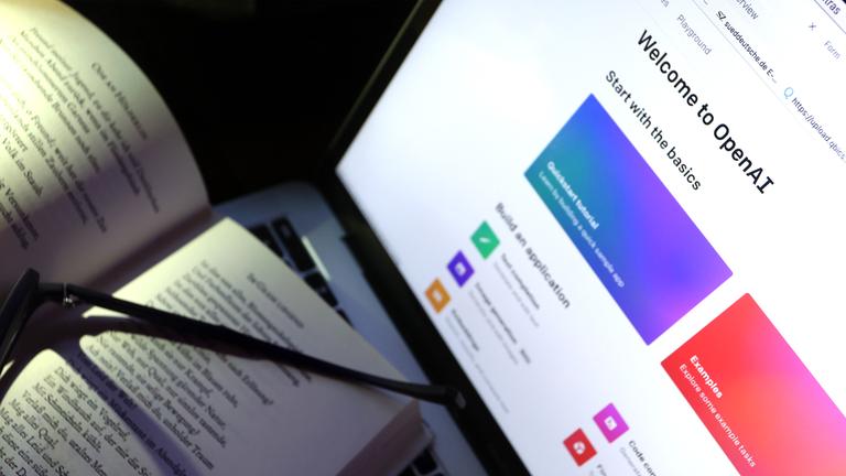 Ein Buch liegt an einem Bildschirm auf dem die Homepage von ChatGPT aufgerufen ist. (Symbolbild)