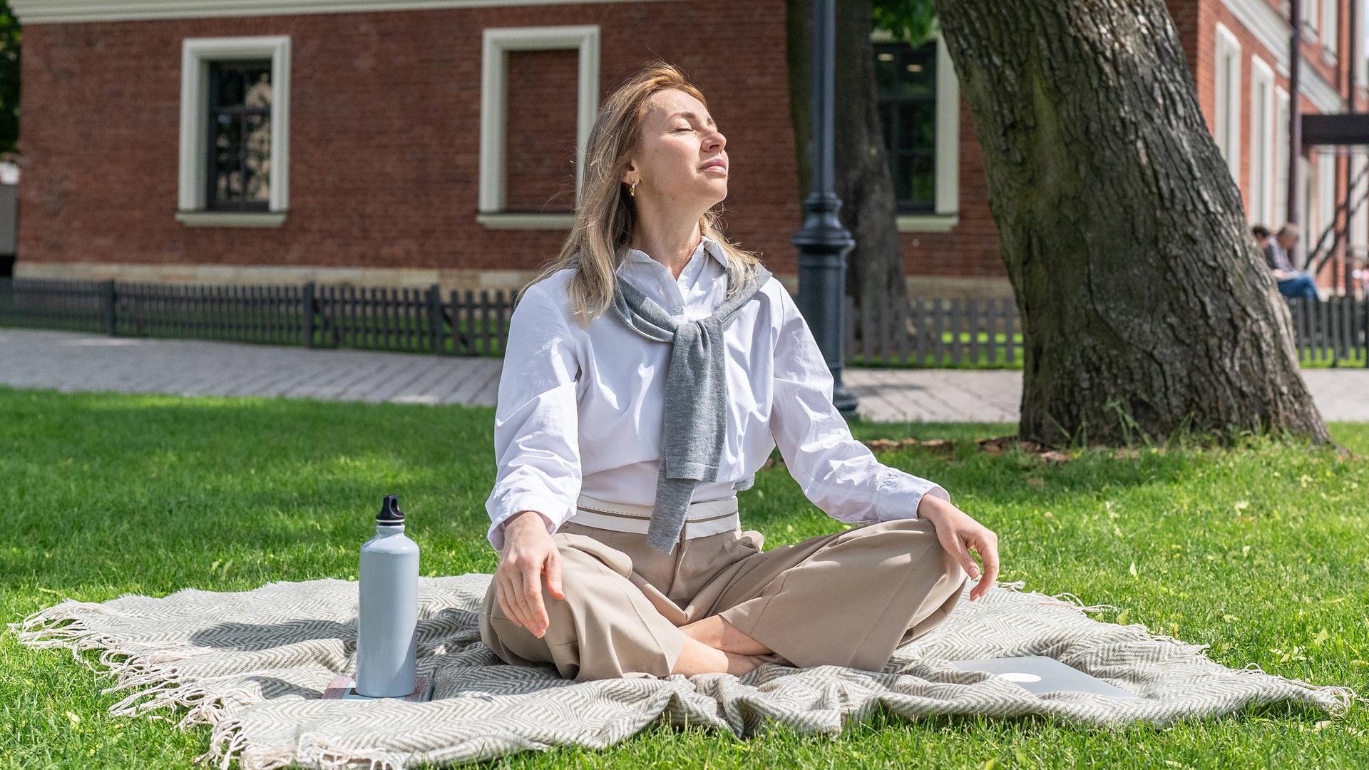 Eine Frau sitzt entspannt auf dem Rasen in einem grünen Park und genießt die warme Sommersonne. (Symbolbild)