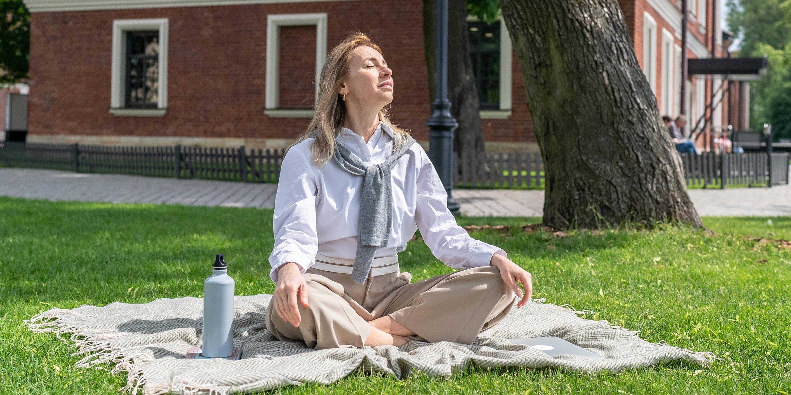 Eine Frau sitzt entspannt auf dem Rasen in einem grünen Park und genießt die warme Sommersonne. (Symbolbild)