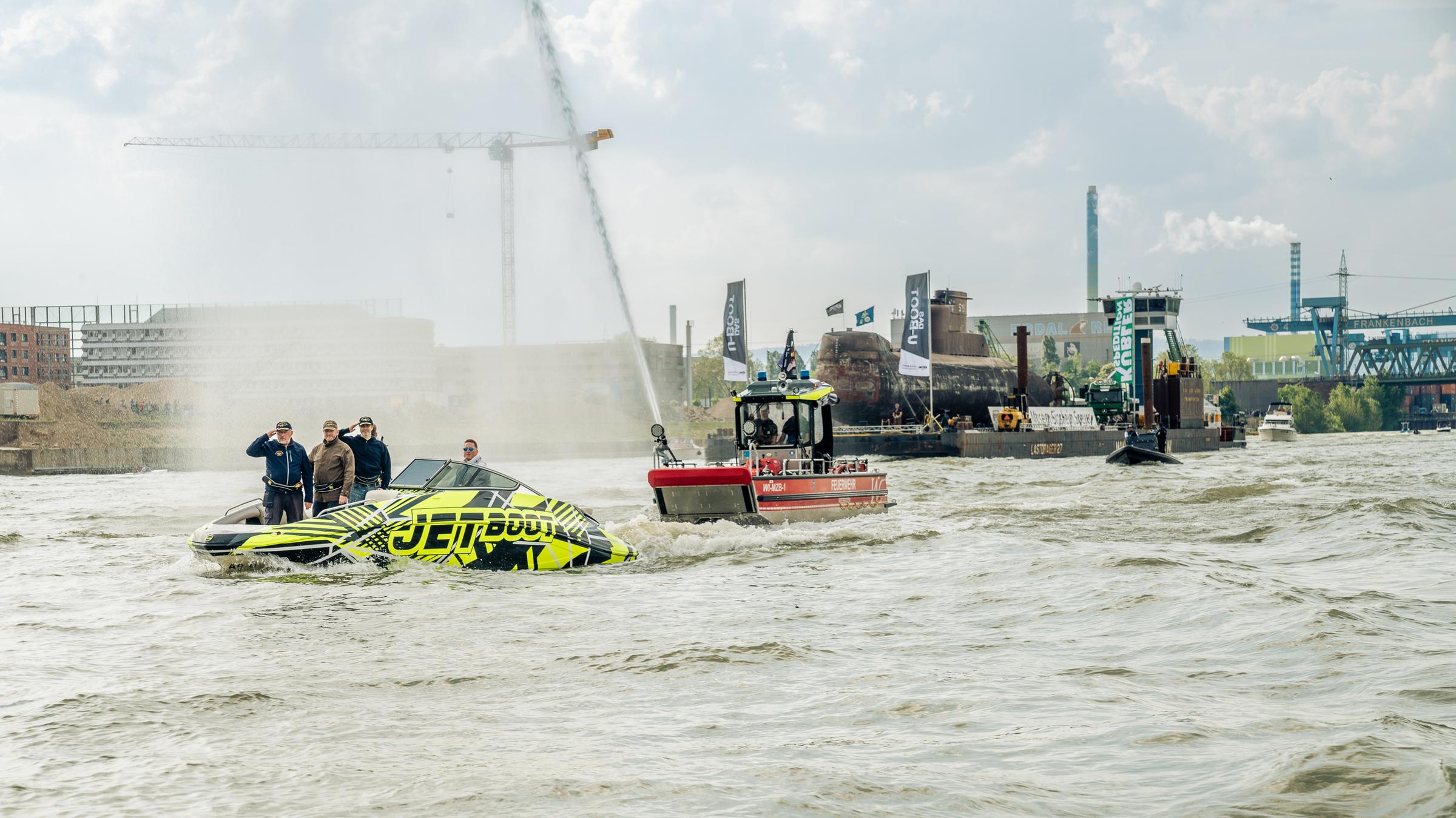 Das U-Boot wird von einem Schlepper gezogen, Menschen stehen auf einem Beiboot, darüber eine Wasserfontäne.
