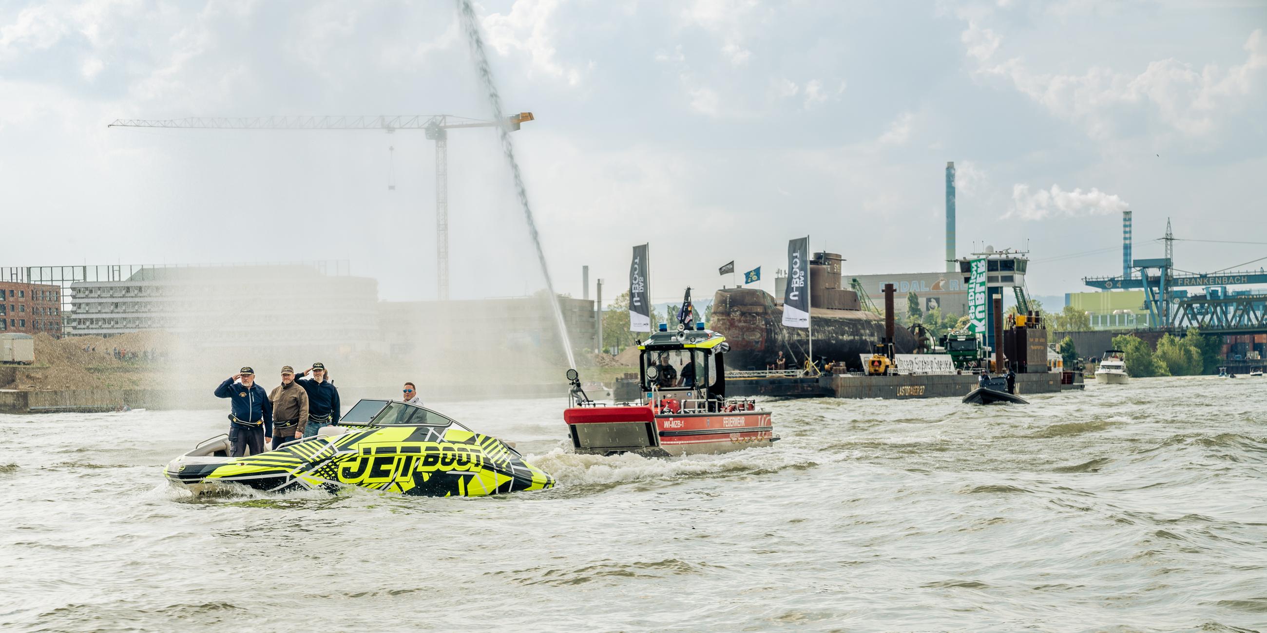 Das U-Boot wird von einem Schlepper gezogen, Menschen stehen auf einem Beiboot, darüber eine Wasserfontäne.