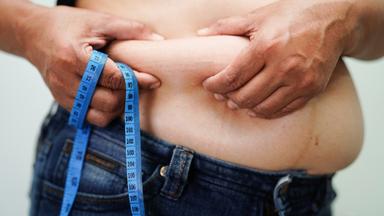 Eine übergewichtige Frau hält sich ihren Bauch und hat ein Maßband in der Hand.