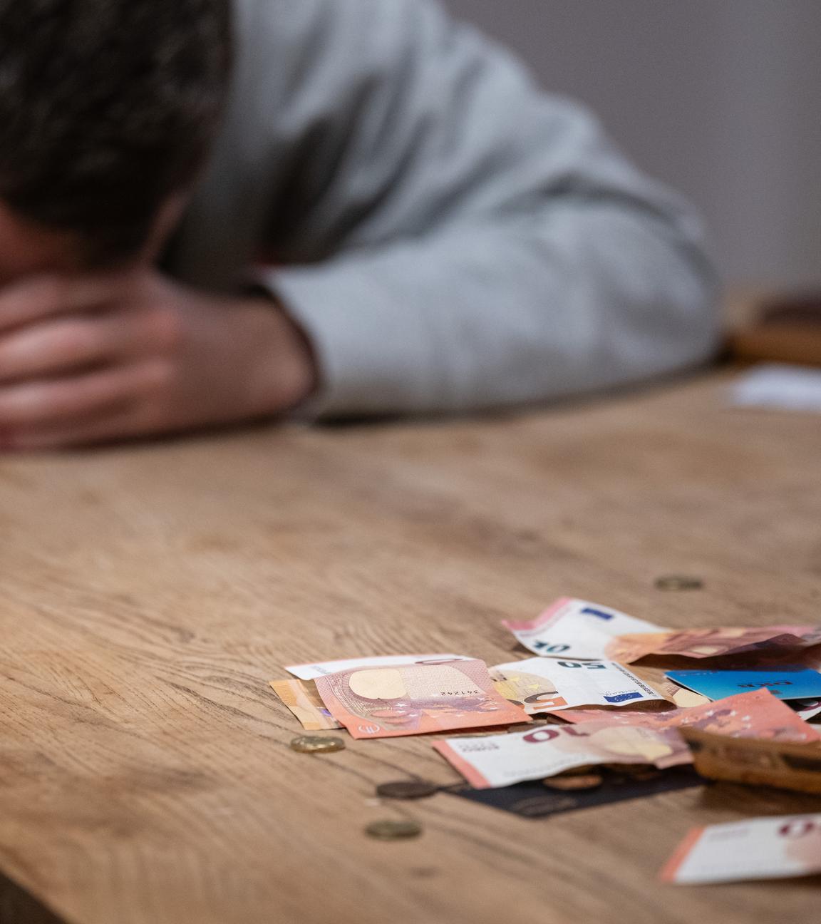 Ein Mann hat seinen Kopf auf seine Hände gelegt, während sich vor ihm Geldscheine, Münzen und Bankkarten auf einem Tisch befinden. 