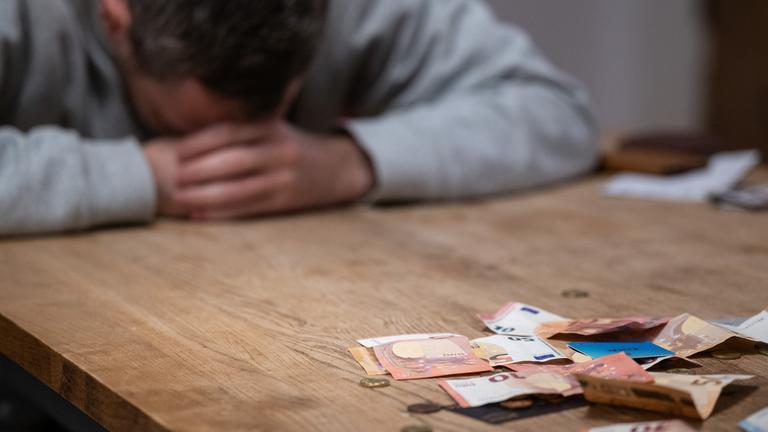 Ein Mann hat seinen Kopf auf seine Hände gelegt, während sich vor ihm Geldscheine, Münzen und Bankkarten auf einem Tisch befinden. 