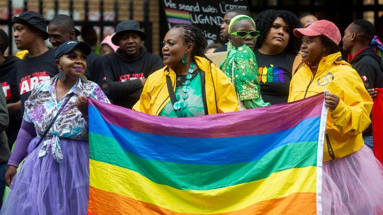 Bunt gekleidete Menschen auf einem Protest. Im Mittelpunkt steht eine Frau, die eine Regenbogenflagge hält.