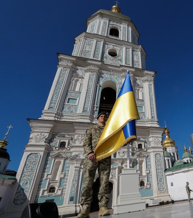 Soldat trägt ukrainische Flagge vor einer Kathedrale in Kiew. Bild von Bodenhöhe nach oben aufgenommen.