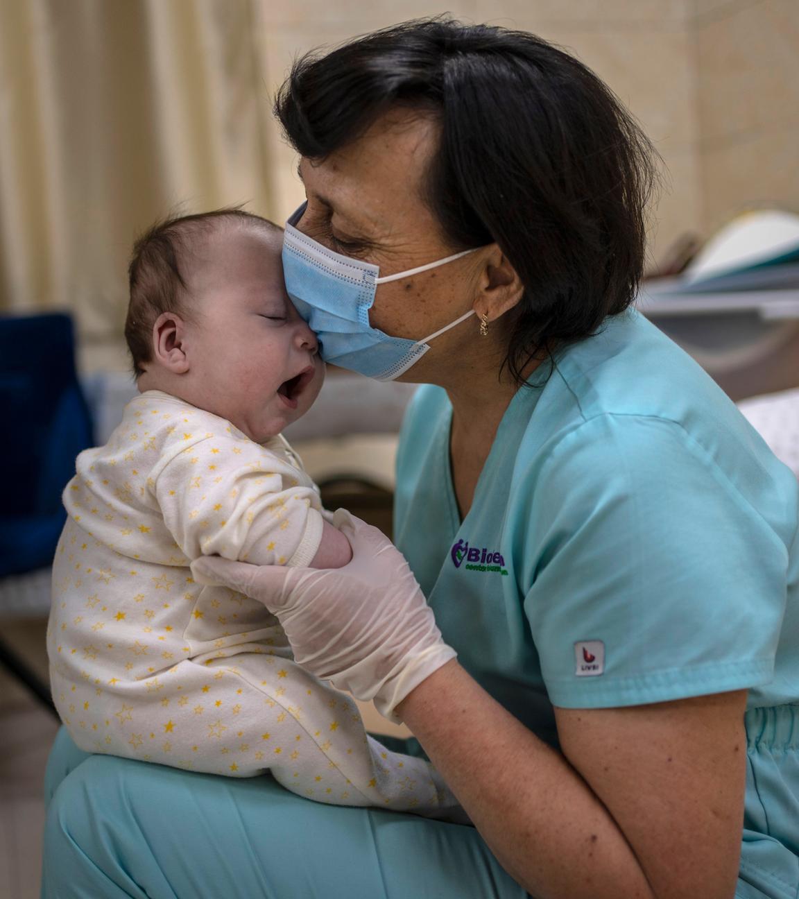 Eine Frau mit einem Mund-Nasen-Schutz hält ein Baby auf ihrem Schoß