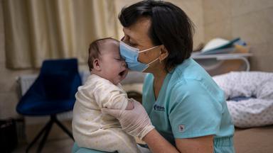 Eine Frau mit einem Mund-Nasen-Schutz hält ein Baby auf ihrem Schoß