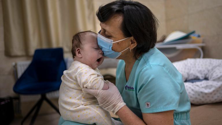 Eine Frau mit einem Mund-Nasen-Schutz hält ein Baby auf ihrem Schoß