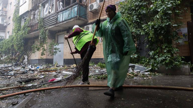 Zwei Personen beseitigen Schäden nach Angriff in Charkiw, eine Person trägt eine gelbe Weste.