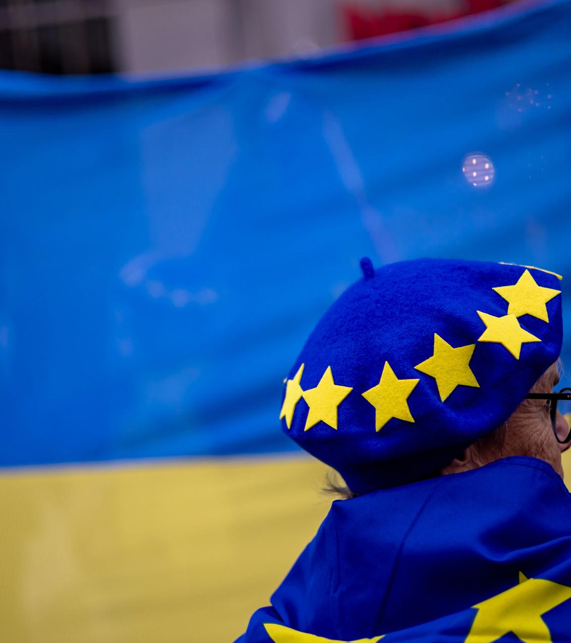 Eine Frau mit Hut und Flagge mit EU-Symbolen steht vor einer großen ukrainischen Flagge