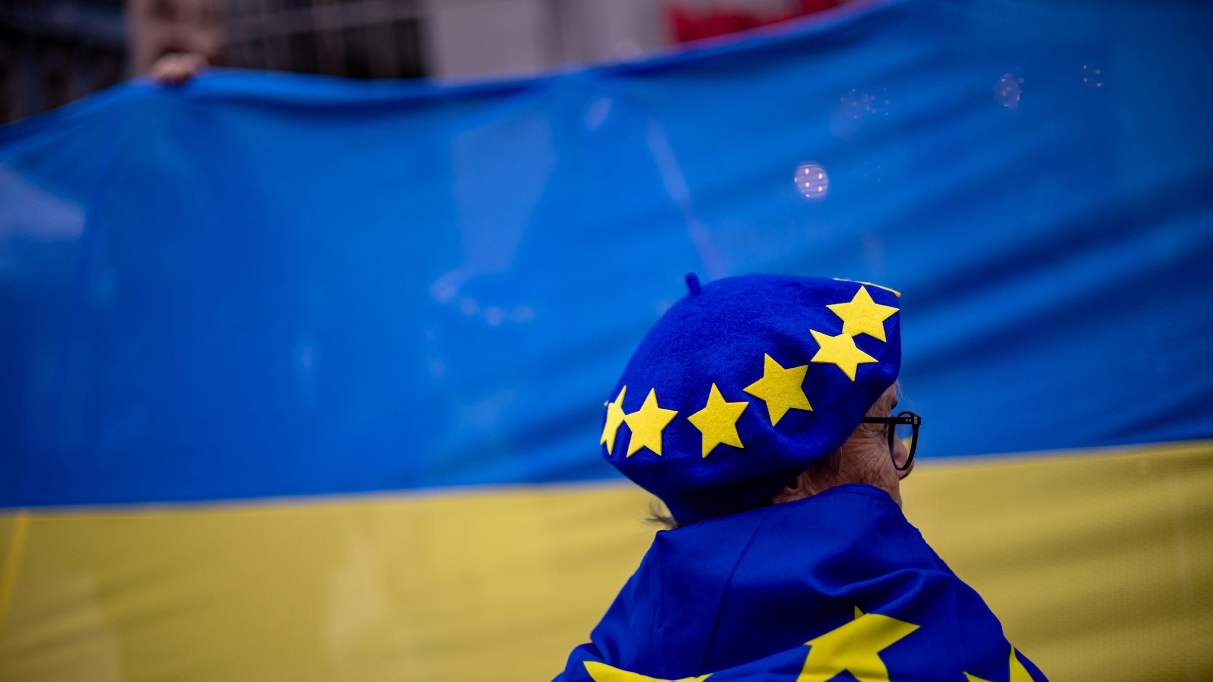 Eine Frau mit Hut und Flagge mit EU-Symbolen steht vor einer großen ukrainischen Flagge