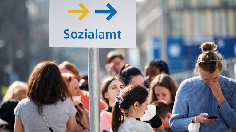 Flüchtlinge, darunter der größte Teil aus der Ukraine, und eine Helferin (r) stehen vor einer Außenstelle des Sachgebiets Flüchtlinge der Ausländerbehörden und der Abteilung Flüchtlinge des Sozialamts in der Innenstadt in einer Schlange