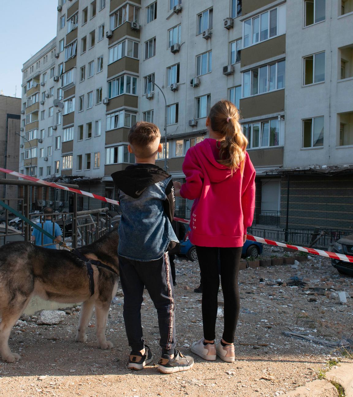 Zwei Kinder blicken auf beschädigte Gebäude und Trümmer. Sie sind von hinten fotografiert. Neben ihnen steht ein Hund