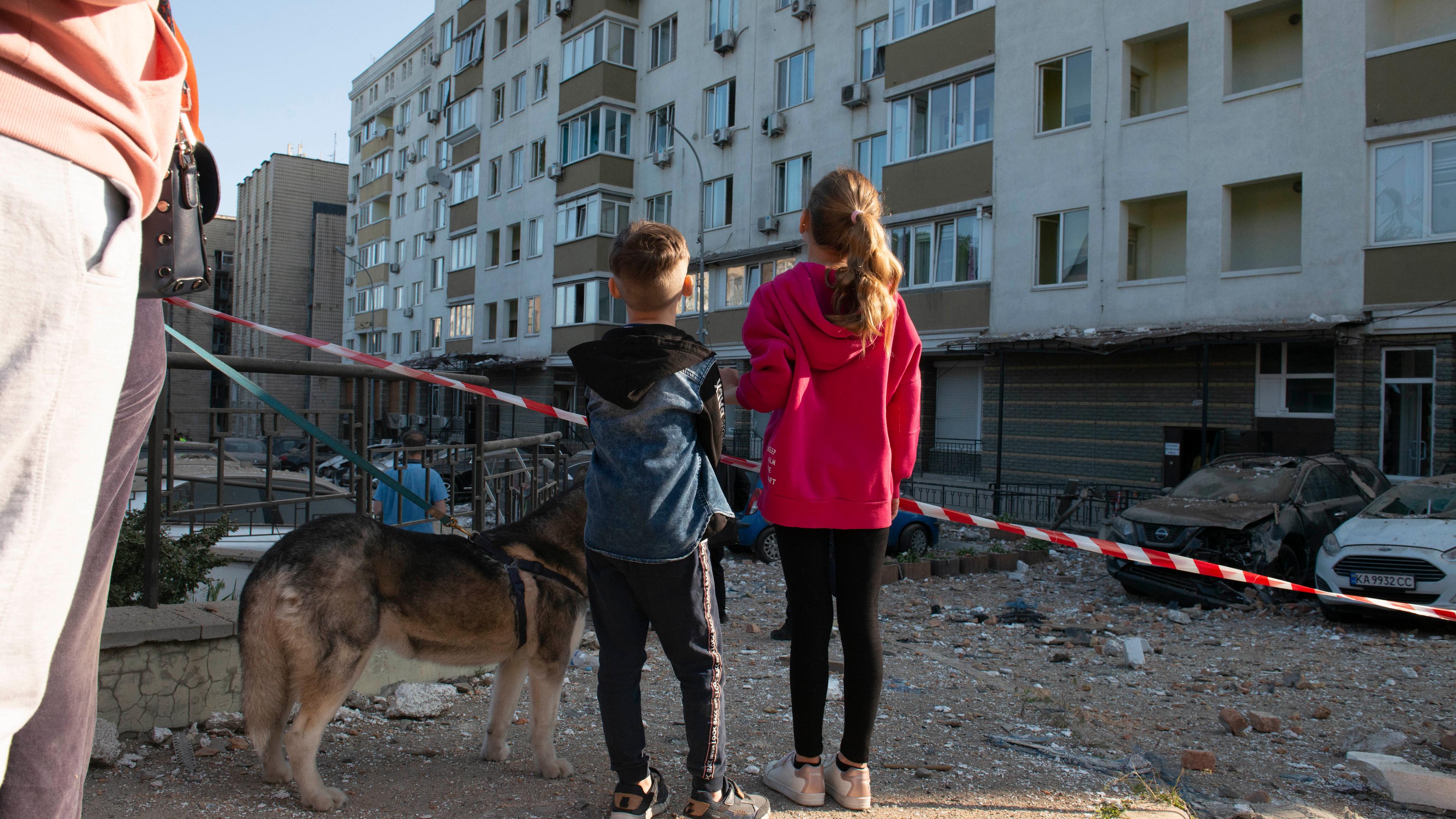 Zwei Kinder blicken auf beschädigte Gebäude und Trümmer. Sie sind von hinten fotografiert. Neben ihnen steht ein Hund