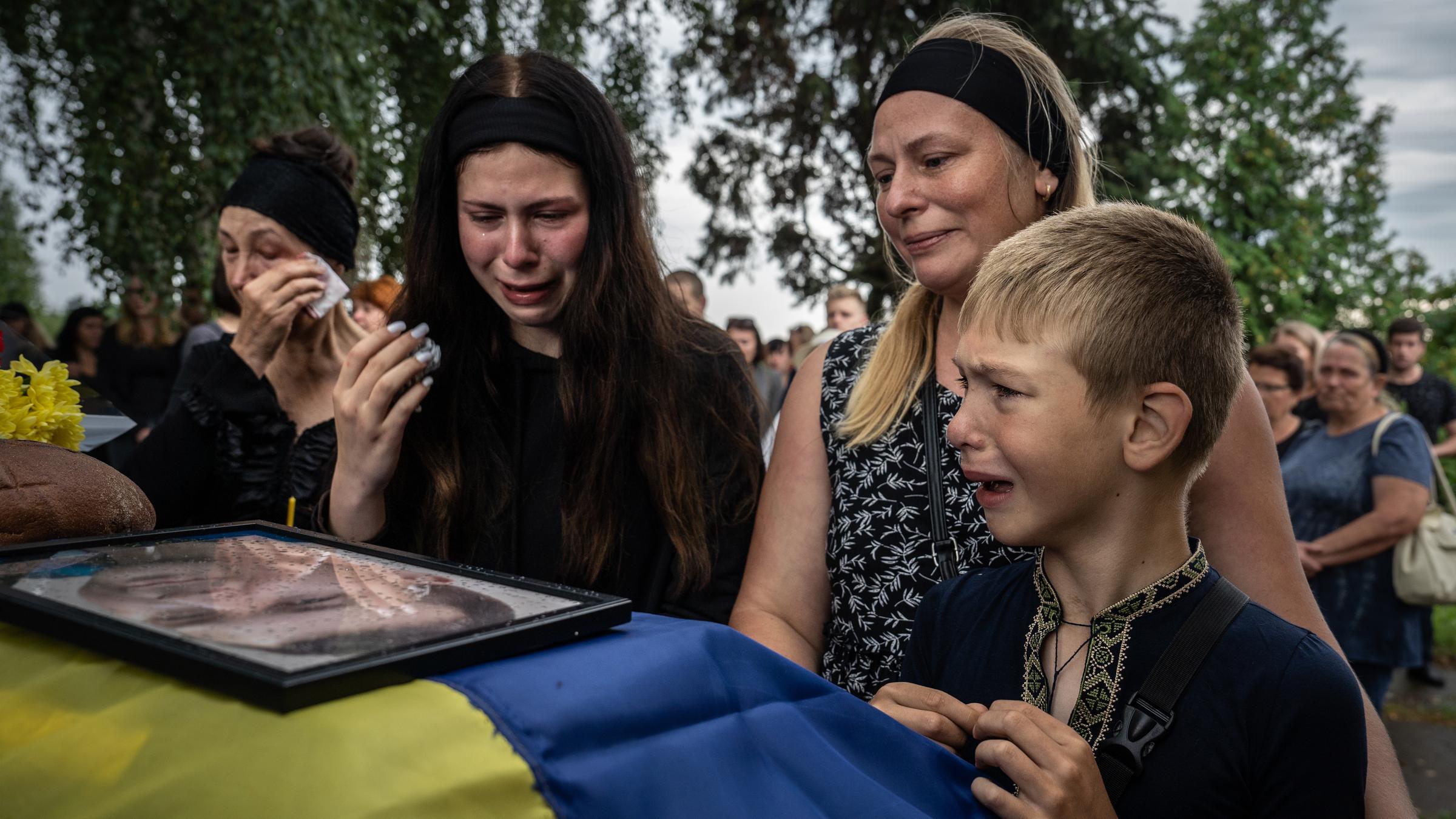 Zwei Frauen und ein kleiner Junge stehen um einen Sarg, auf dem die ukrainische Flagge und ein gerahmtes Bild liegen.