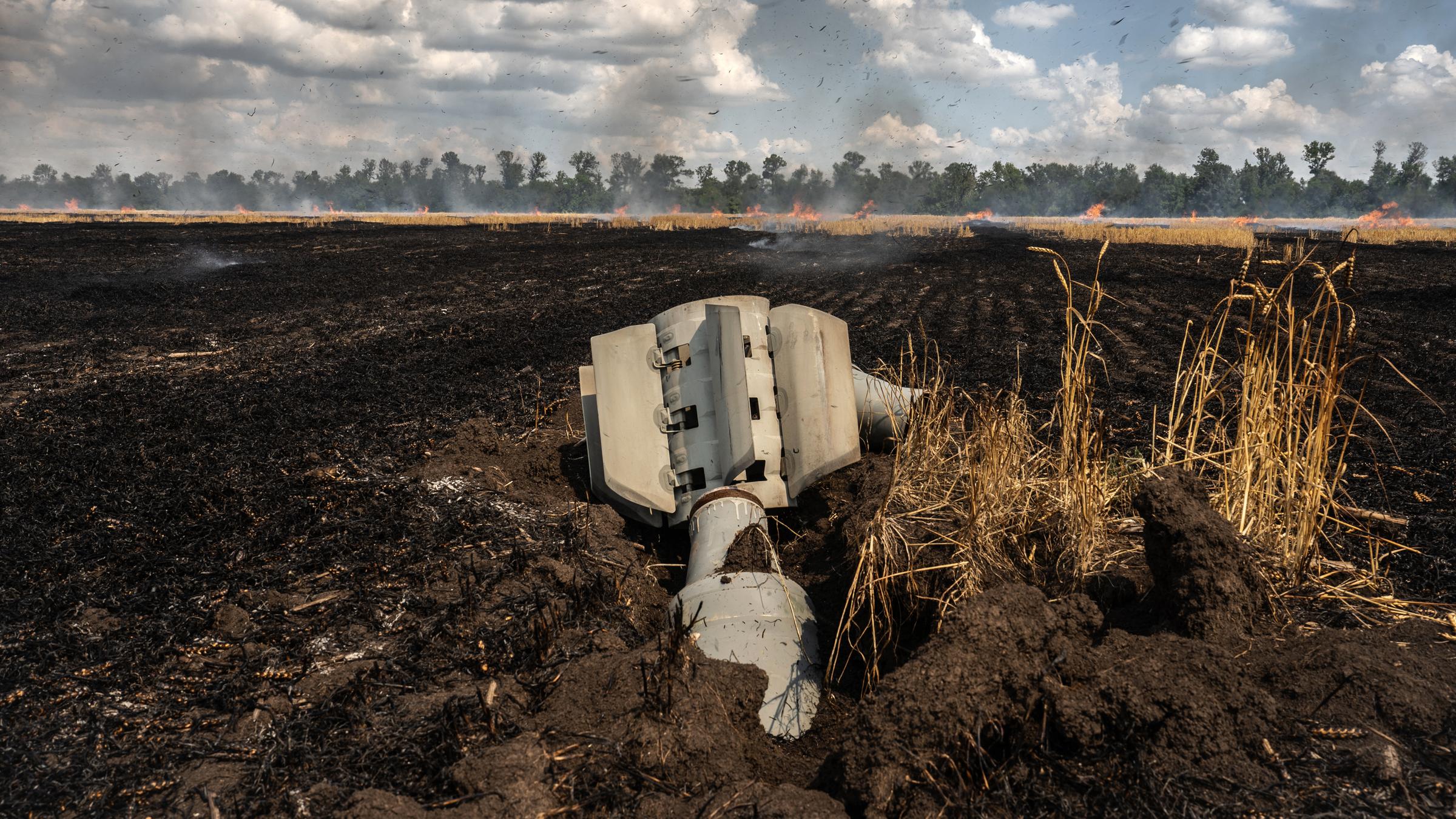 Eine Rakete liegt auf einem Weizenfeld, im Hintergrund ist ein Feuer zu sehen.