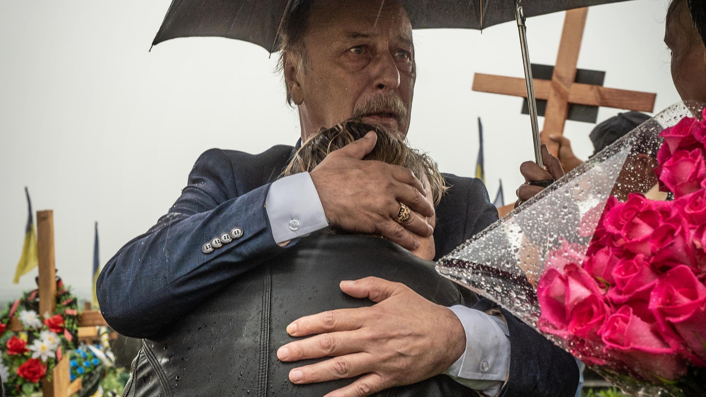 Ein Mann hält eine Frau in den Armen, über ihnen ein Regenschirm, im Hintergrund ist ein Friedhof zu sehen.