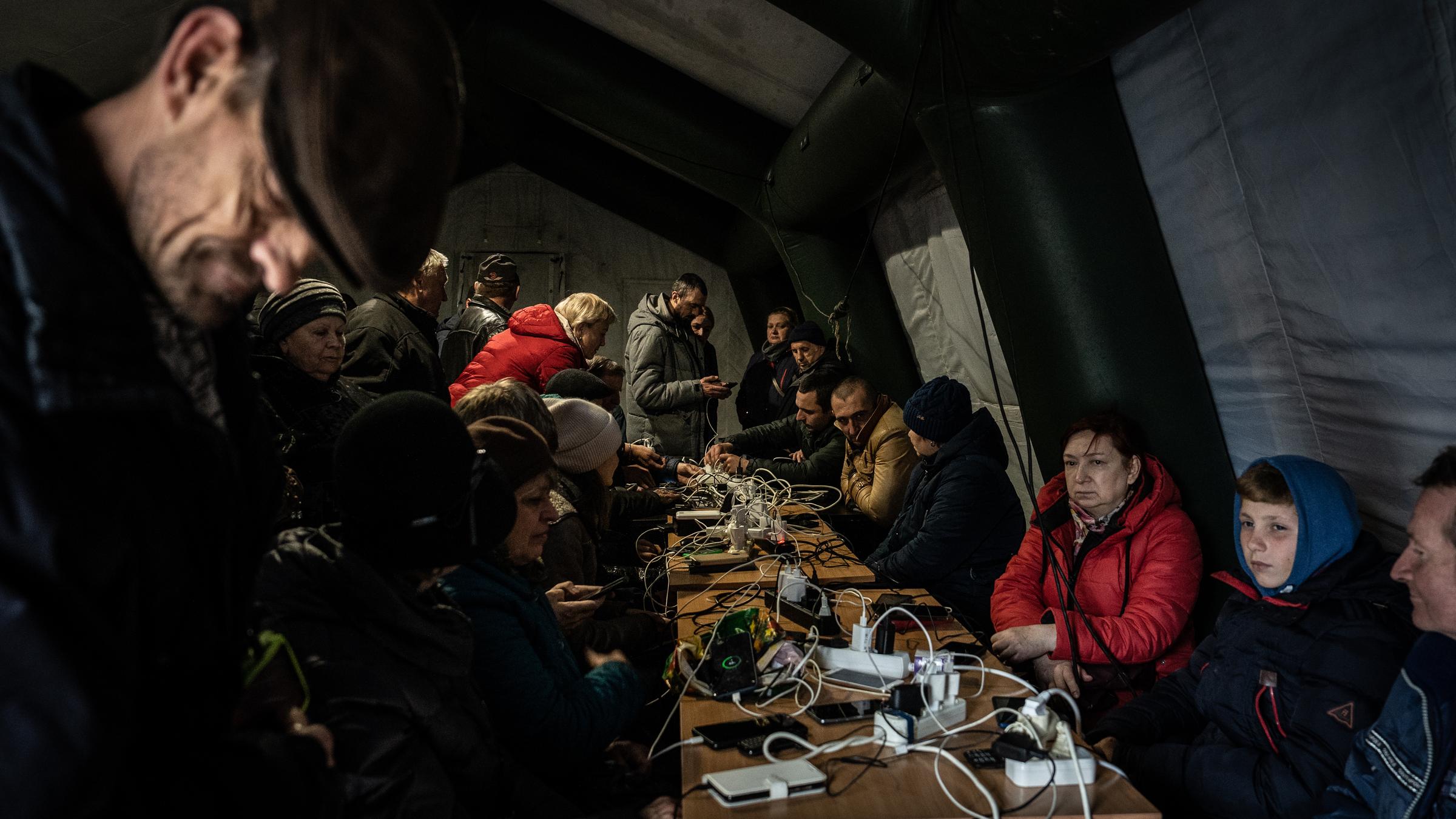 Das Bild zeigt Menschen in einem Notzelt, die um einen Tisch herumsitzen und ihre Geräte laden.