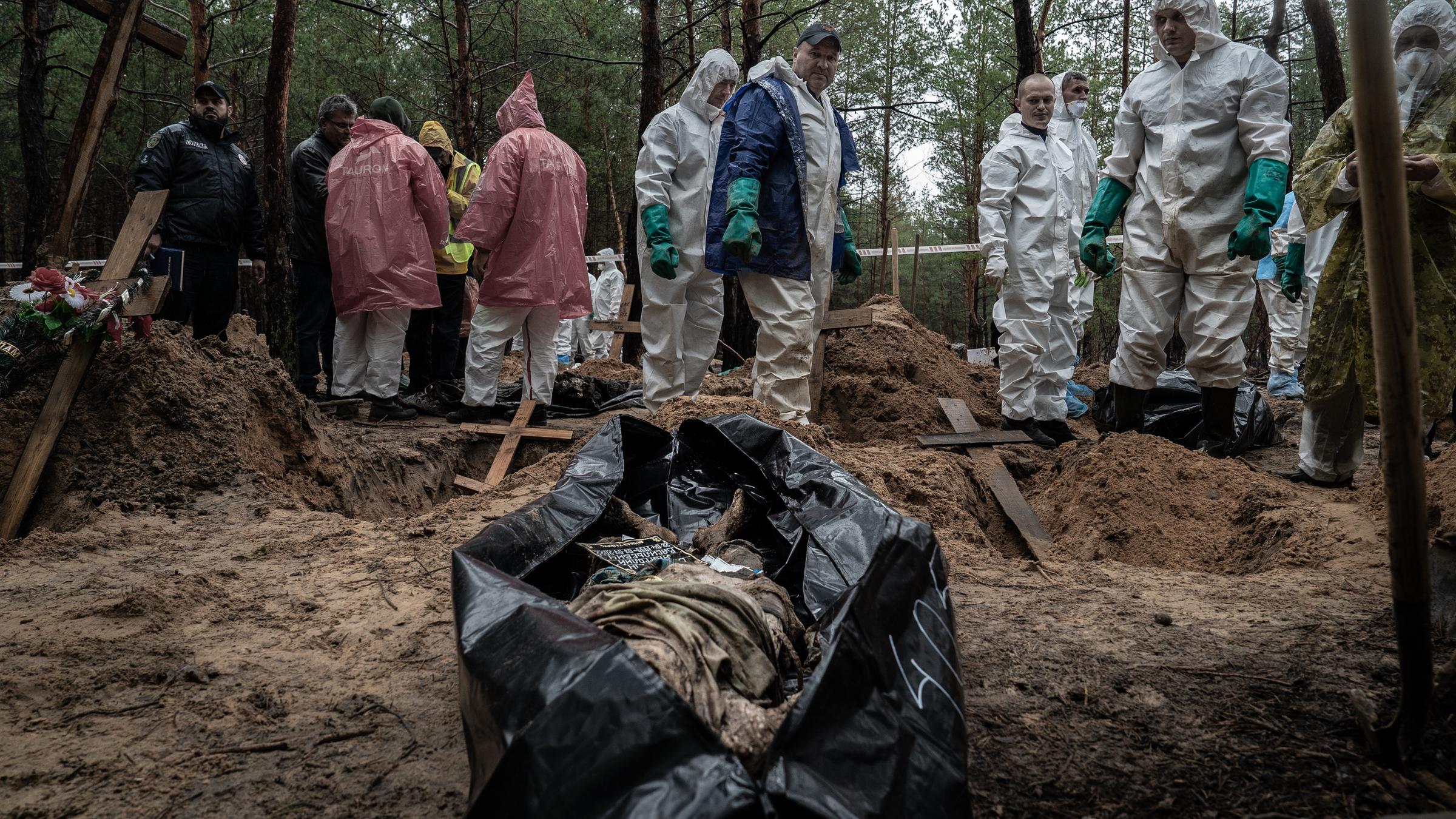 Menschen in Plastikanzügen stehen um einen Leichensack im Wald.