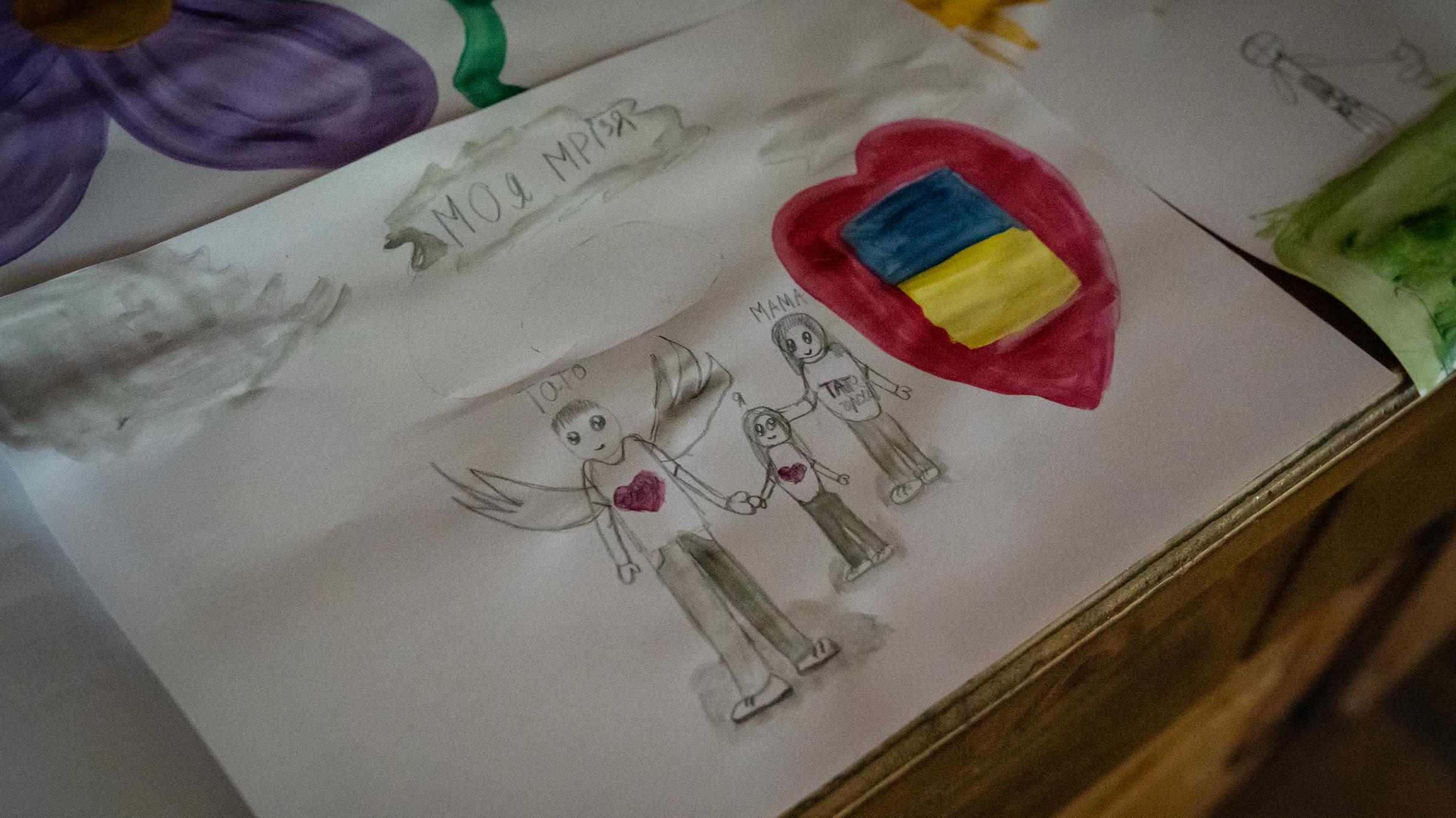 Selbstgemaltes Bild auf einem Blatt Papier, das drei Menschen und die ukrainische Flagge zeigt.