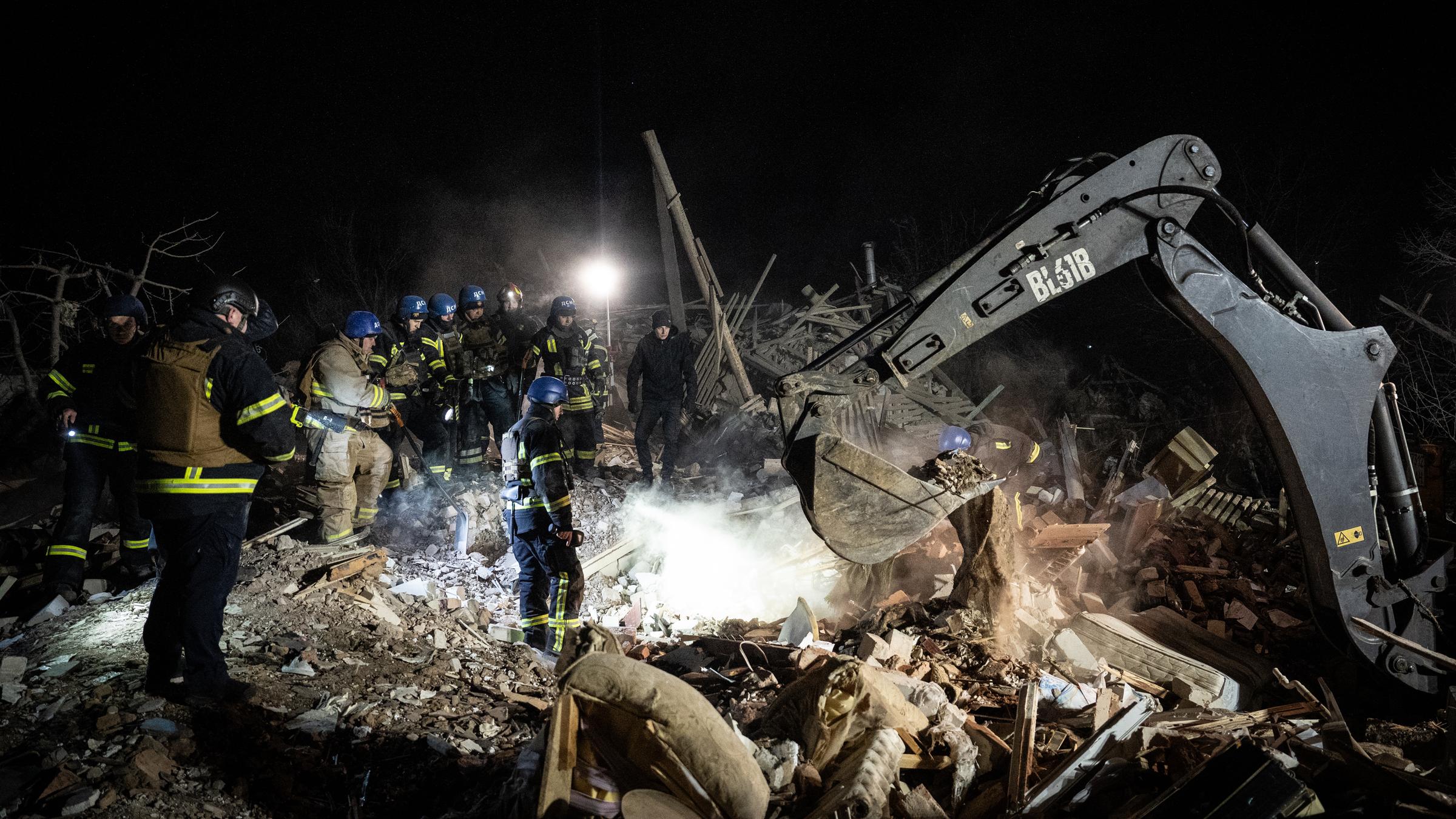 Rettungskräfte stehen in Trümmern, neben ihnen gräbt ein Bagger.