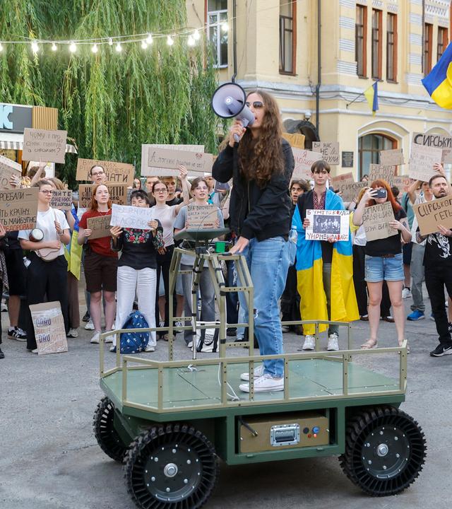 Ukrainer bedienen ein unbemanntes Bodenfahrzeug (UGV) und halten Schilder mit den Aufschriften „Fedorov ist der Verteidigungsminister“, „Ändert nichts, was funktioniert“, „Finger weg von Fedorov“ und „Holt Fedorov zurück“ in die Höhe.