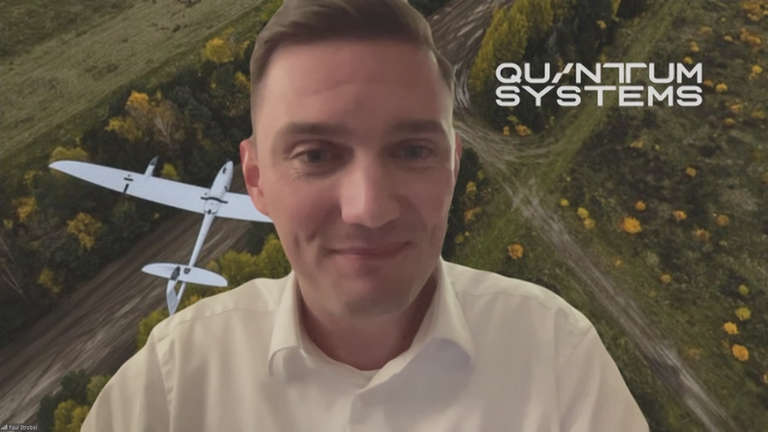 Quantum-Systems Sprecher Paul Strobel vor Grafik einer Landschaft mit Drohne.