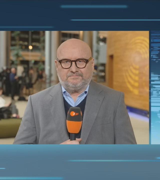 Eine geteilte Bildschirmansicht zeigt links Ulf Röller in Brüssel und rechts Moderator Christopher Wehrmann im Nachrichtenstudio. Röller trägt eine dunkle, runde Brille, ein graues Sakko und einen dunkelblauen Pullover über einem hellblauen Hemd. Vor seiner Brust hält er ein orange-schwarzes ZDF-Mikrofon. Hinter ihm sind unscharf mehrere Menschen in einem großen Innenraum zu sehen. Wehrmann trägt ein braunes, kariertes Sakko, ein hellblaues Hemd und eine beigefarbene Krawatte. Hinter ihm erscheint eine hellblaue Weltkarte auf blauem Grund. Thema der Schalte sind die EU und die Arktis.