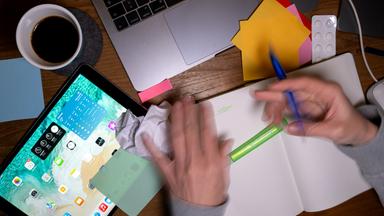 Ein unaufgeräumter Schreibtisch mit einem Tablet, einer Kaffetasse, einem Notizbuch und verschwommenen Händen.