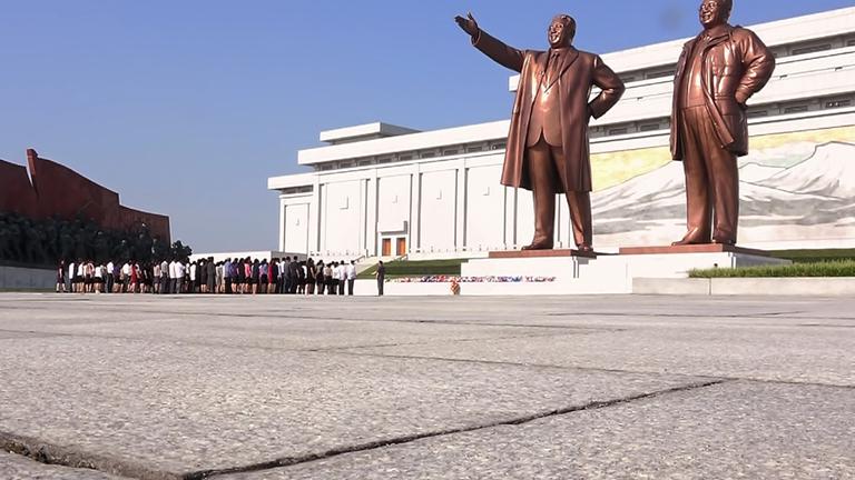 "Undercover in Nordkorea - Im Reich des Kim Jong Un": Zwei monumentale Bronzestatuen der verstorbenen koreanischen Führer stehen vor blauen Himmel. Davor steht eine Gruppe von Menschen.