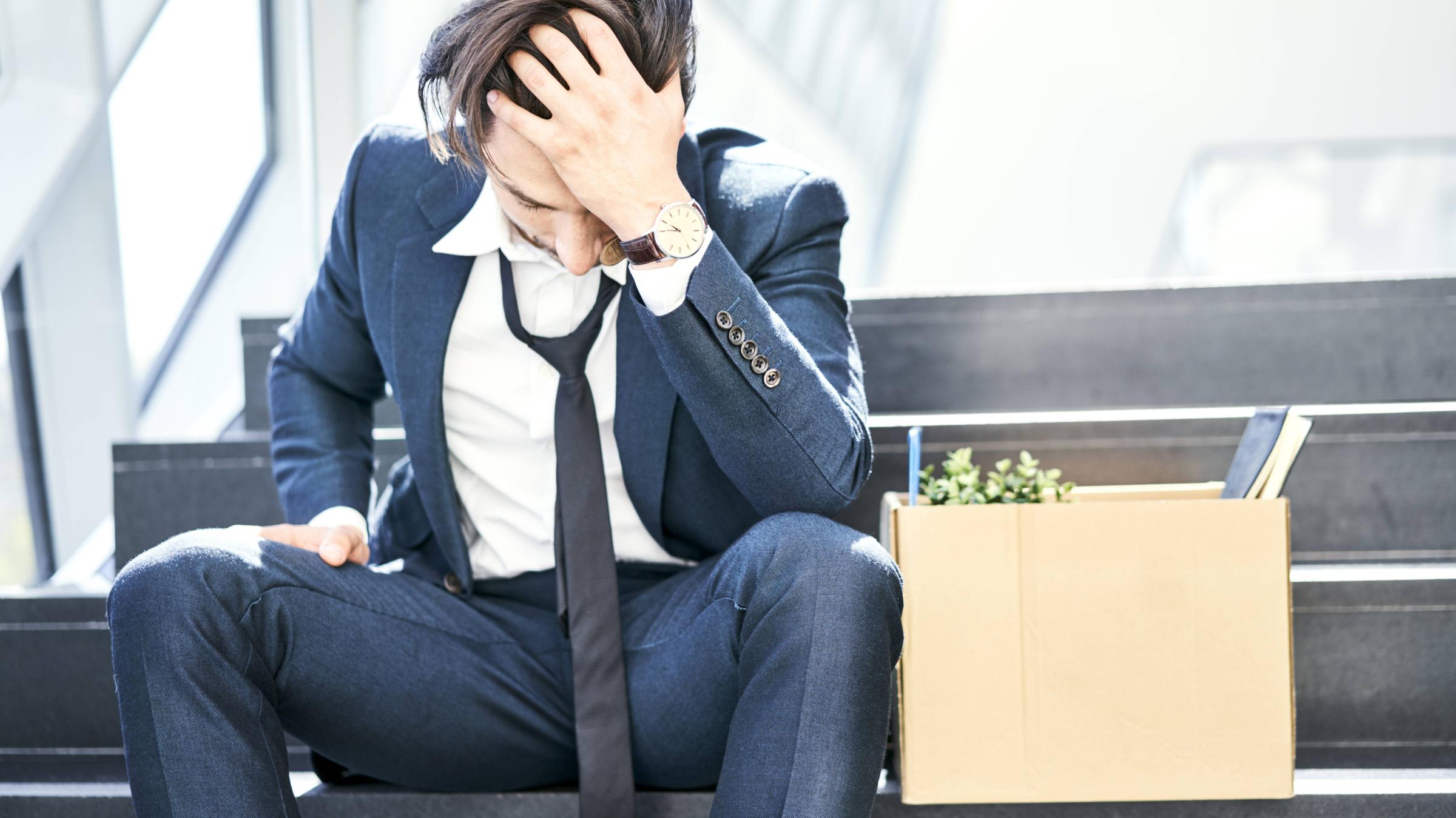 Ein Mann in professioneller Kleidung sitzt verweifelt neben einem Karton voller Büromaterial und einer kleinen Pflanze.