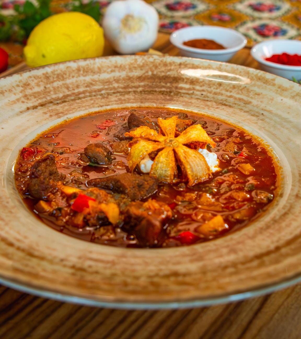 Eine ungarische Suppe mit einer frittierten Zwiebelblüte in einem braunen Suppenteller. Im Hintergrund stehen allerlei Gewürze in kleinen Schälchen.