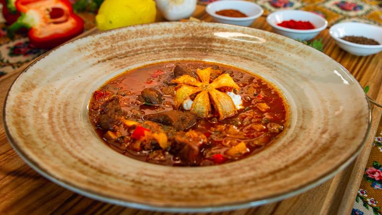 Eine ungarische Suppe mit einer frittierten Zwiebelblüte in einem braunen Suppenteller. Im Hintergrund stehen allerlei Gewürze in kleinen Schälchen.