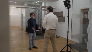 Peter Techet spricht mit einer Person in weißem Hemd, die der Kamera den Rücken zugewandt hat.