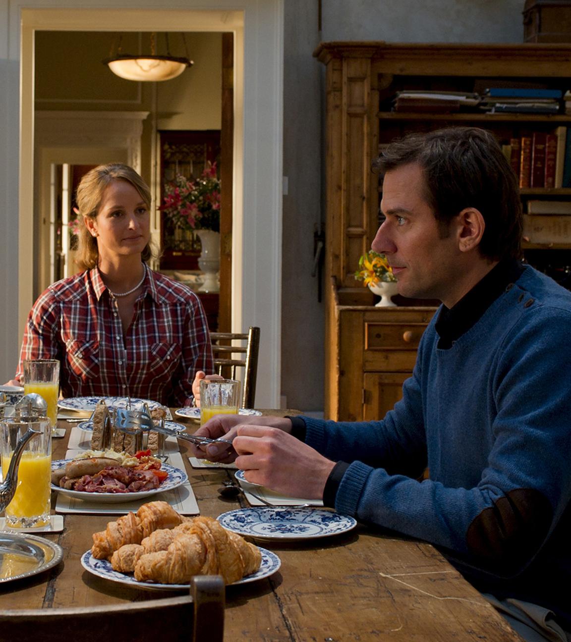 Gillian (Mitte, Hintergrund) sitzt zusammen mit Dylan (rechts, Vordergrund) und dessen Mutter Lady Ava Brooks (links, Vordergrund) am Frühstückstisch.