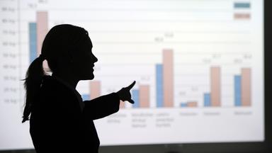 Eine frau steht in einem Büro und erklärt eine an die Wand projizierte Grafik.