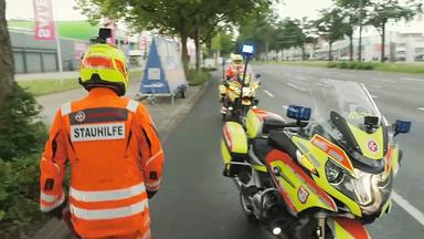 Eine Einsatzkraft in orangefarbener Schutzkleidung steht neben zwei Motorrädern am Straßenrand. Auf der Jacke ist der Schriftzug ‚Stauhilfe‘ zu sehen.