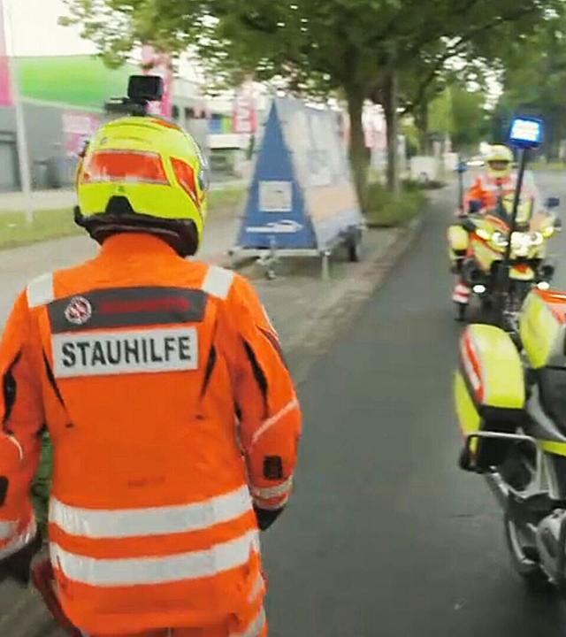 Eine Einsatzkraft in orangefarbener Schutzkleidung steht neben zwei Motorrädern am Straßenrand. Auf der Jacke ist der Schriftzug ‚Stauhilfe‘ zu sehen.