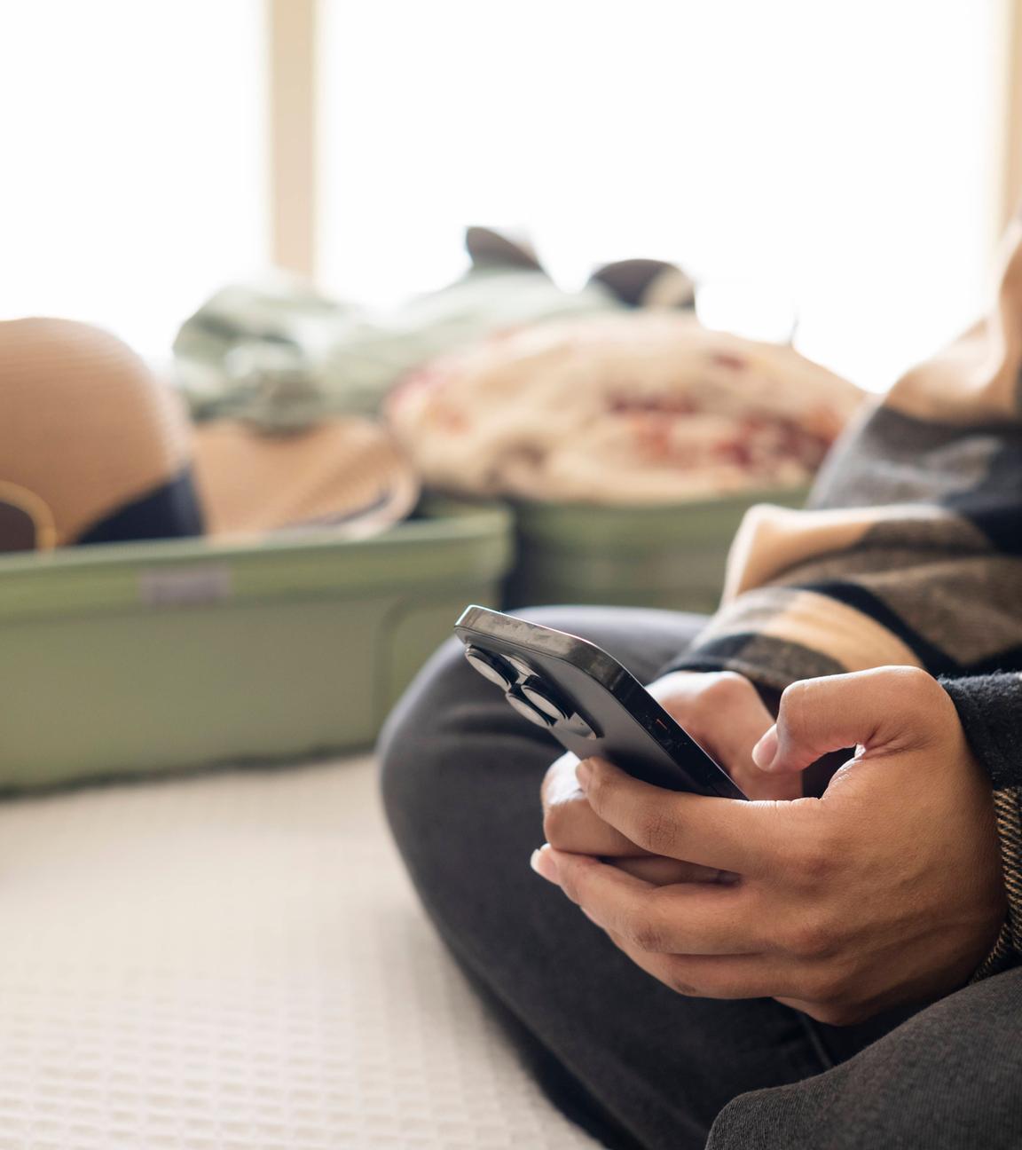 Frau bucht eine Reise auf dem Smartphone, im Hinergrund ein gepackter Koffer.