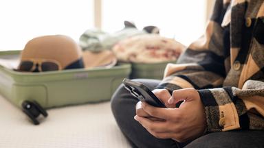 Frau bucht eine Reise auf dem Smartphone, im Hinergrund ein gepackter Koffer.