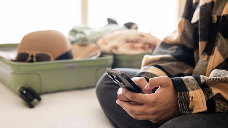 Frau bucht eine Reise auf dem Smartphone, im Hinergrund ein gepackter Koffer.