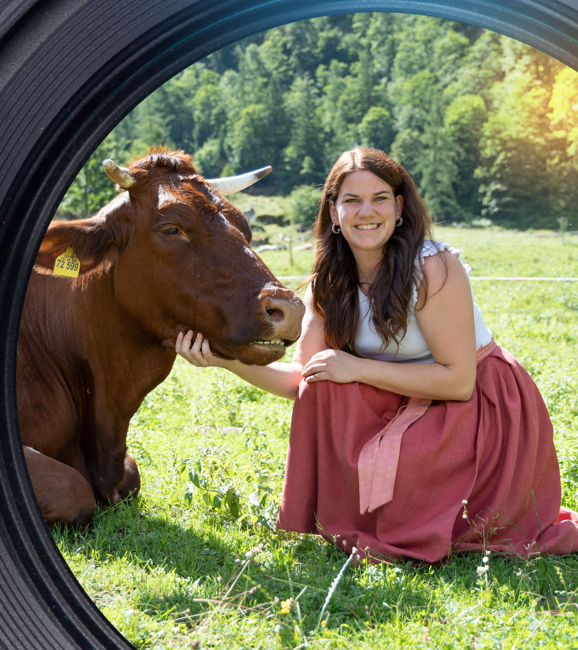 Veronika hockt neben einer liegenden Kuh auf einer Weide und schaut lächelnd in die Kamera.