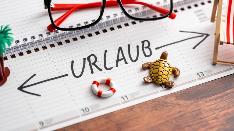 Schrift "Urlaub" auf einem Kalender mit Markierung der ganzen Woche. Liegestuhl und Strandzubehör auf Kalender.